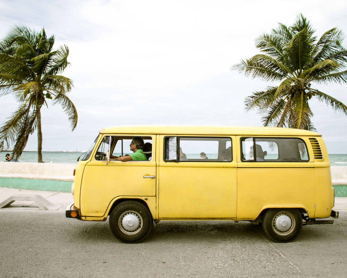 photo of an old, mustard yellow van on a road adjacent to s beach. Palm trees and the ocean are in view in the back.