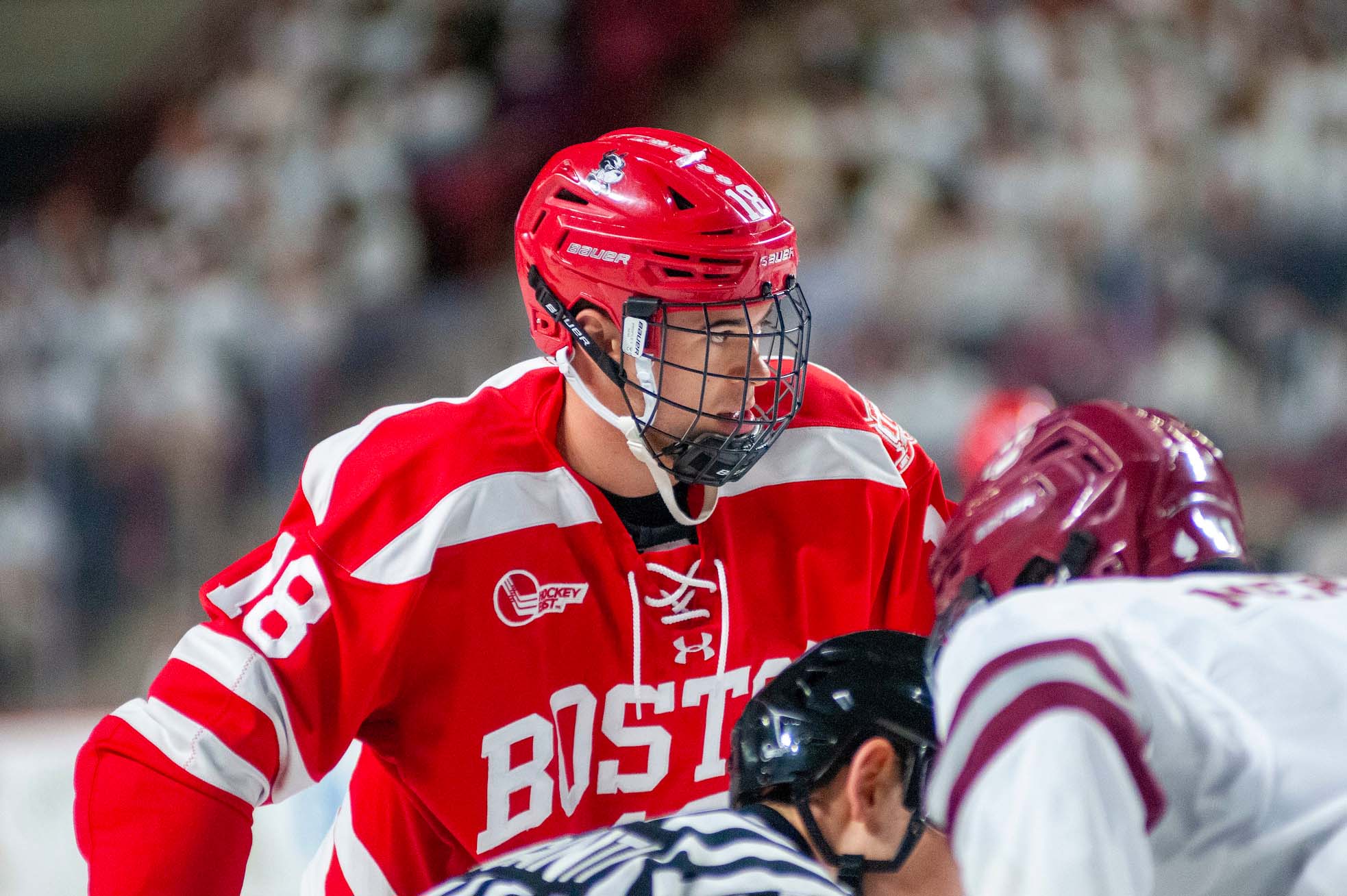 action shot of BU men's hockey player Jay O’Brien, a white man, at face off. The referee stands between O'Brien and the opposing UMass player.
