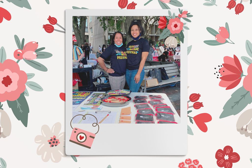 Image: Polaroid style photo of 2 women of color, masks lowered and smiling, as they stand and pose together in front of a table with merchandise for their club. Polaroid is placed on a tan background with a pattern of red, tan,  and pink flowers, red hearts, and a pink camera