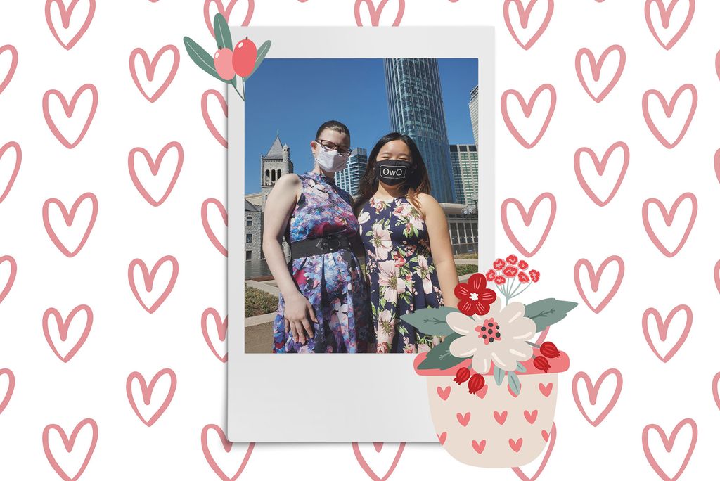 Image: Polaroid style photo of 2 women wearing face masks and standing in front of a city background as they look and smile towards the camera. Polaroid is placed on a white background with a pattern of pinks hearts. Clipart of a tan vase of red and pink flowers and pink berries border the polaroid.