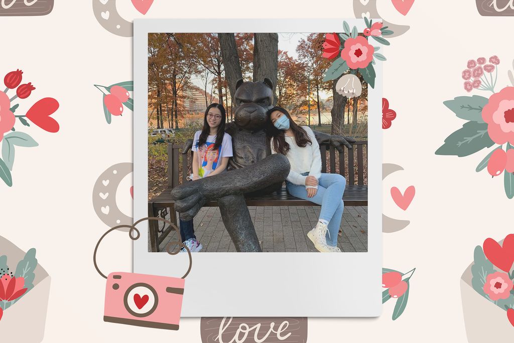 Image: Polaroid style photo of two asian women sitting on either side of a bronze Rhett statue on a bench. One wears a blue mask while the other smiles at the camera. Polaroid is placed on a tan background with a pattern of red and pink flowers, red heart flowers, taupe moons, and a pink camera