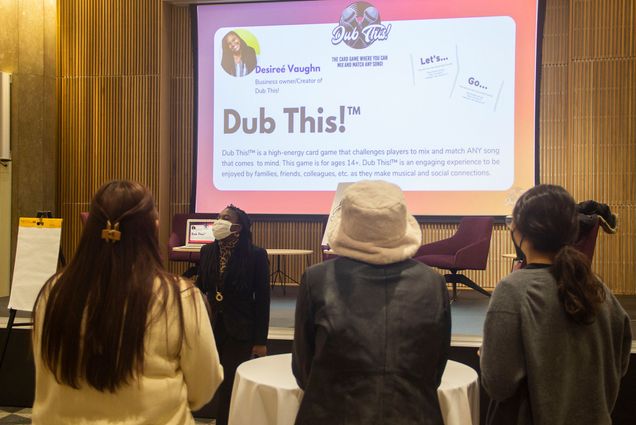 Photo of students facing a large screen as Desireé Vaughn, a black woman wearing a masks stands. Screen reads "Dub This!" and features rules of the game