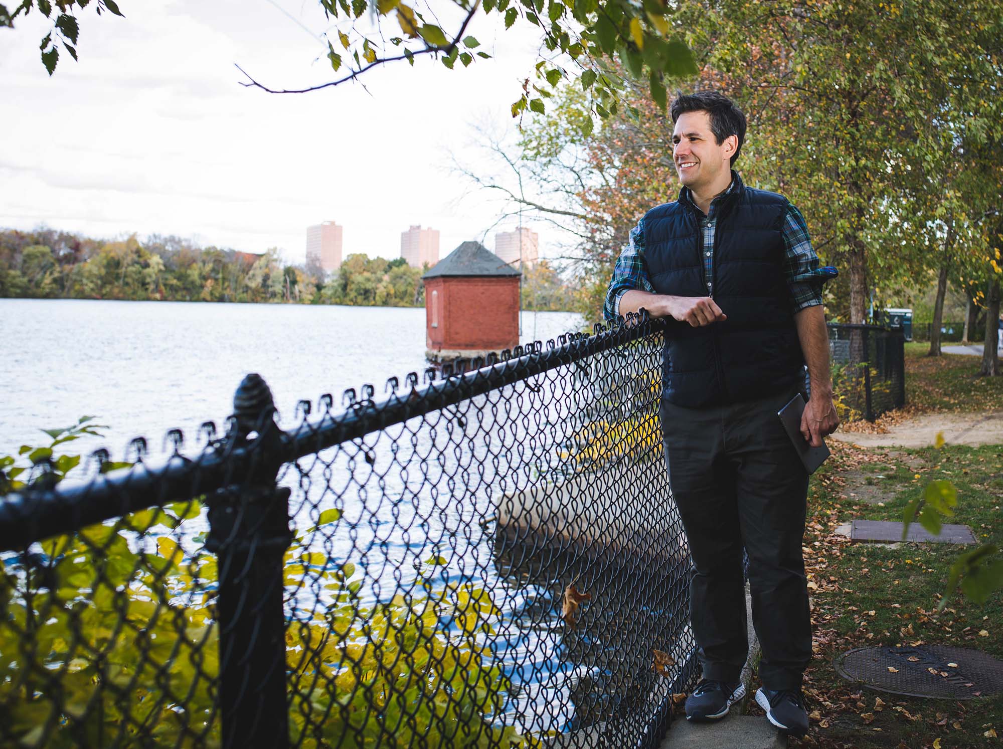 photo of Andrew Robichaud, a white man wearing a blue plaid shirt and a navy puffer vest and dark blue jeans, stands next to a fence and looks out over a river