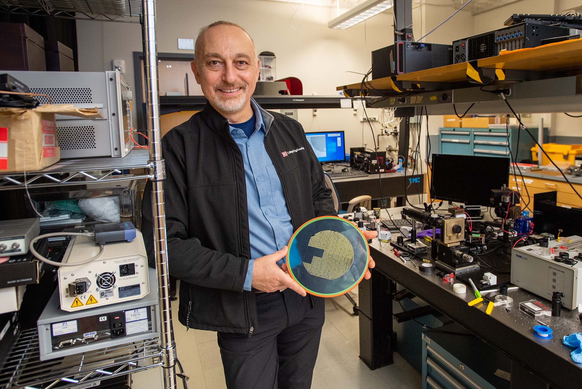 Photo of Selim Ünlü, a tan man wear a blue collared shirt and black jacket, holding a disc-shaped device in his cluttered mechanical engineering lab.