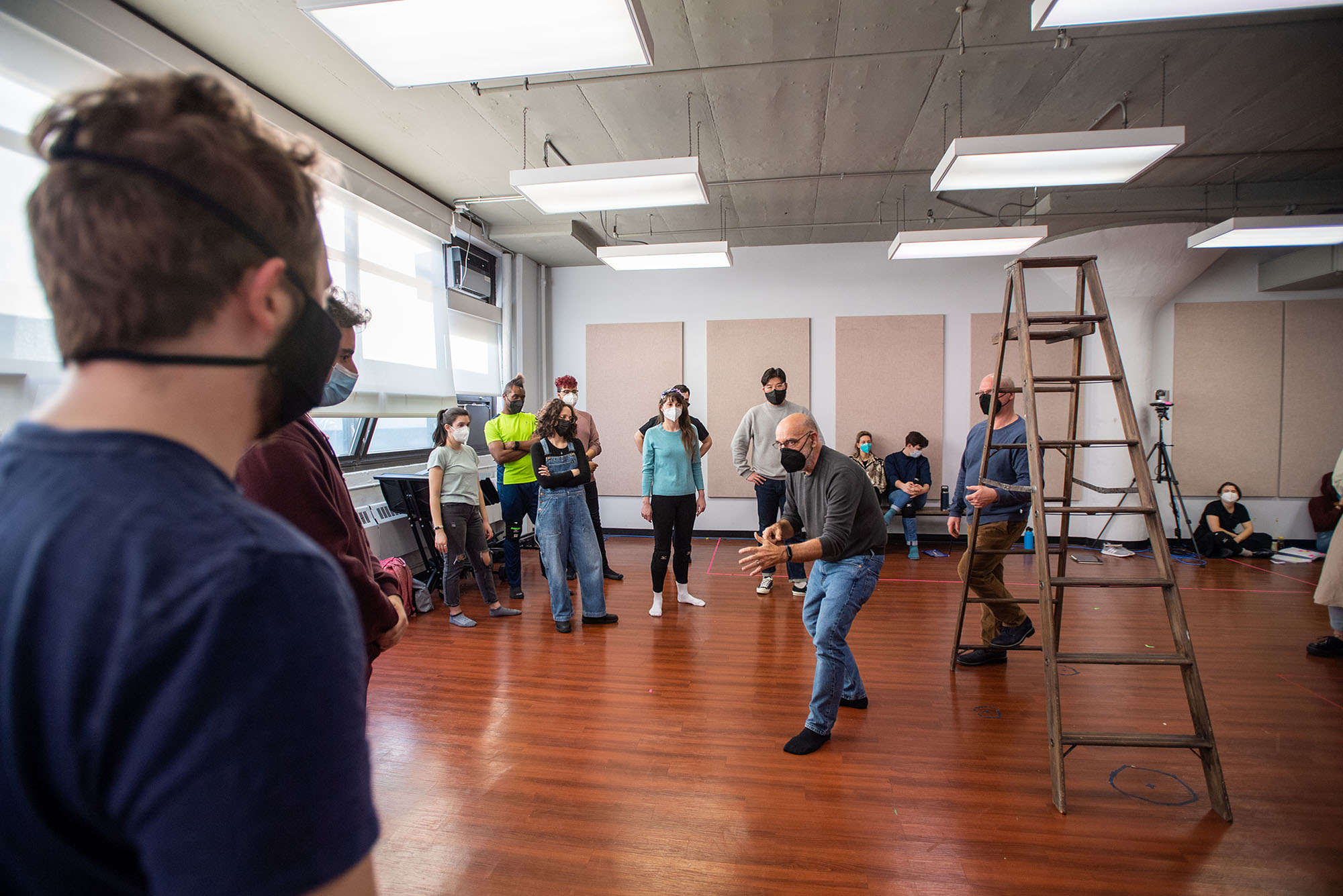 Photo of Stage Director Jim Petosa at the Opera Institute’s rehearsal for "If I Were You". An older, balding white man gestures dramatically in the center of the room next to a ladder as a diverse group of students look on and listen. All are masked.