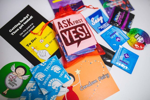 Photo of a variety of different brands of condoms scattered on a white table. Two small postcards included read "Ask first. Yes!" and "Condom Fairy"