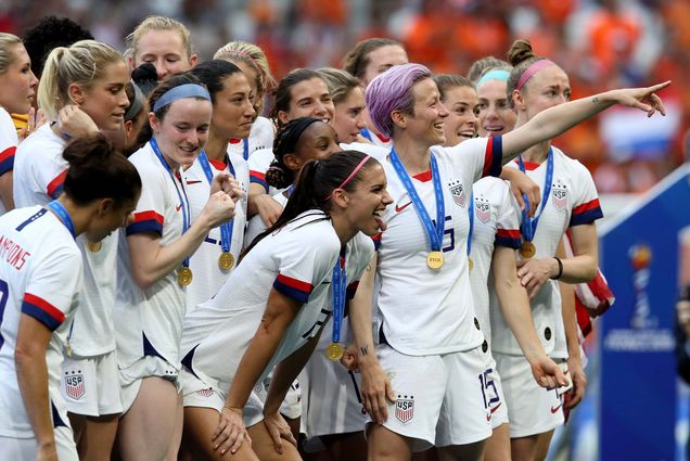 Photo of the US’ national team celebrating with teammates after their 2-0 victory in the 2019 Women’s World Cup final soccer match between United States and the Netherlands at the Stade de Lyon in France, July 7. The women are seen in white jerseys and smile. Many wear colorful headbands and most wear blue medals around their necks.