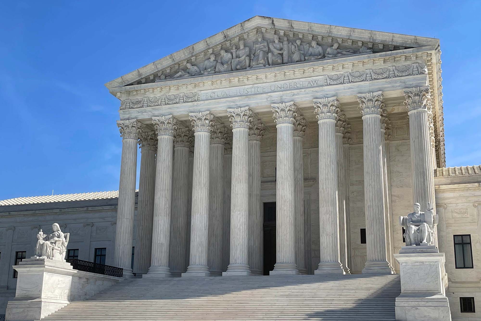 Photo on a sunny day of the exterior of US Supreme Court building. Shows an old, white Roman style building with large pillars and many steps.