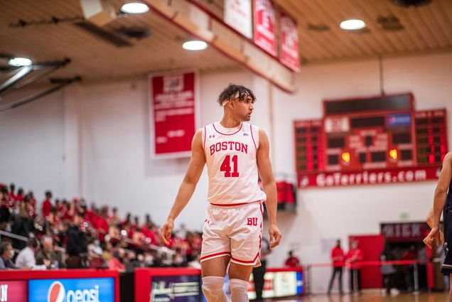 Photo of a BU men's basketball player walking down court. A tan young adult with brown curly hair wears a white and red jersey and looks over his shoulder. Background shows a blurry crowd and scoreboard.