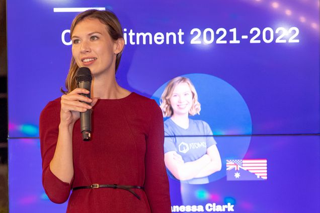Photo of Vanessa Clark, cofounder and CEO of start-up Atomos Space. A white woman with long blonde hair speaks on stage in front of a large projector screen featuring a blue slide that reads "commitment"