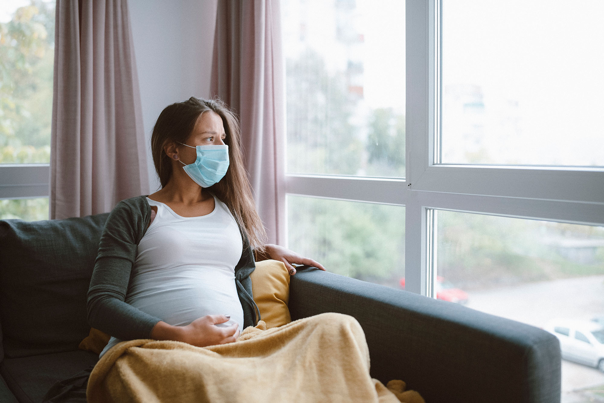 photo of a worried, white pregnant woman sitting at home with protective face mask on and looking through the window on the right.