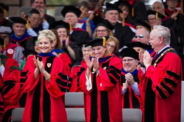 Photo of Ryan Roth Gallo (LAW’99), pictured (left) at the 2018 BU Commencement in full regalia. On the right, Carmen Yulín Cruz Soto (CAS’84) and President Brown are seen. All of them clap, and rows of commencement go-ers in regalia are seen behind them.