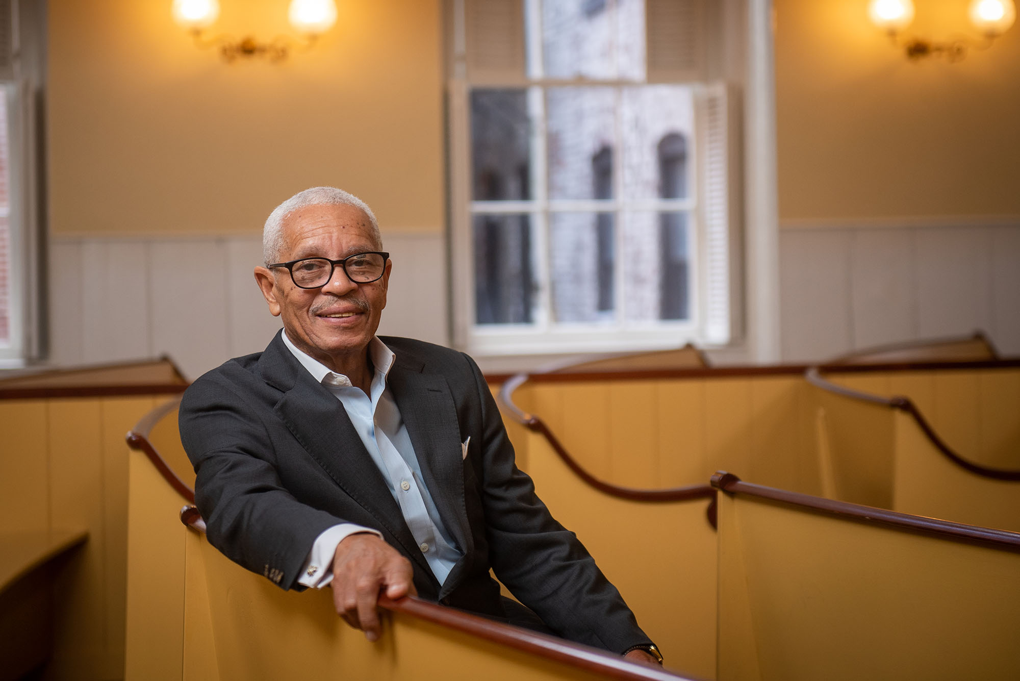 Photo of MET alum Leon Wilson, President and Chief Executive Officer at the Museum of African American History (MAAH) sitting in a pew at the meeting house on November 18, 2021. Wilson is an older Black man with short, graying hair and large black-rimmed glasses, who smiles, wears a suit and rests his right arm on the wooden, painted pews.