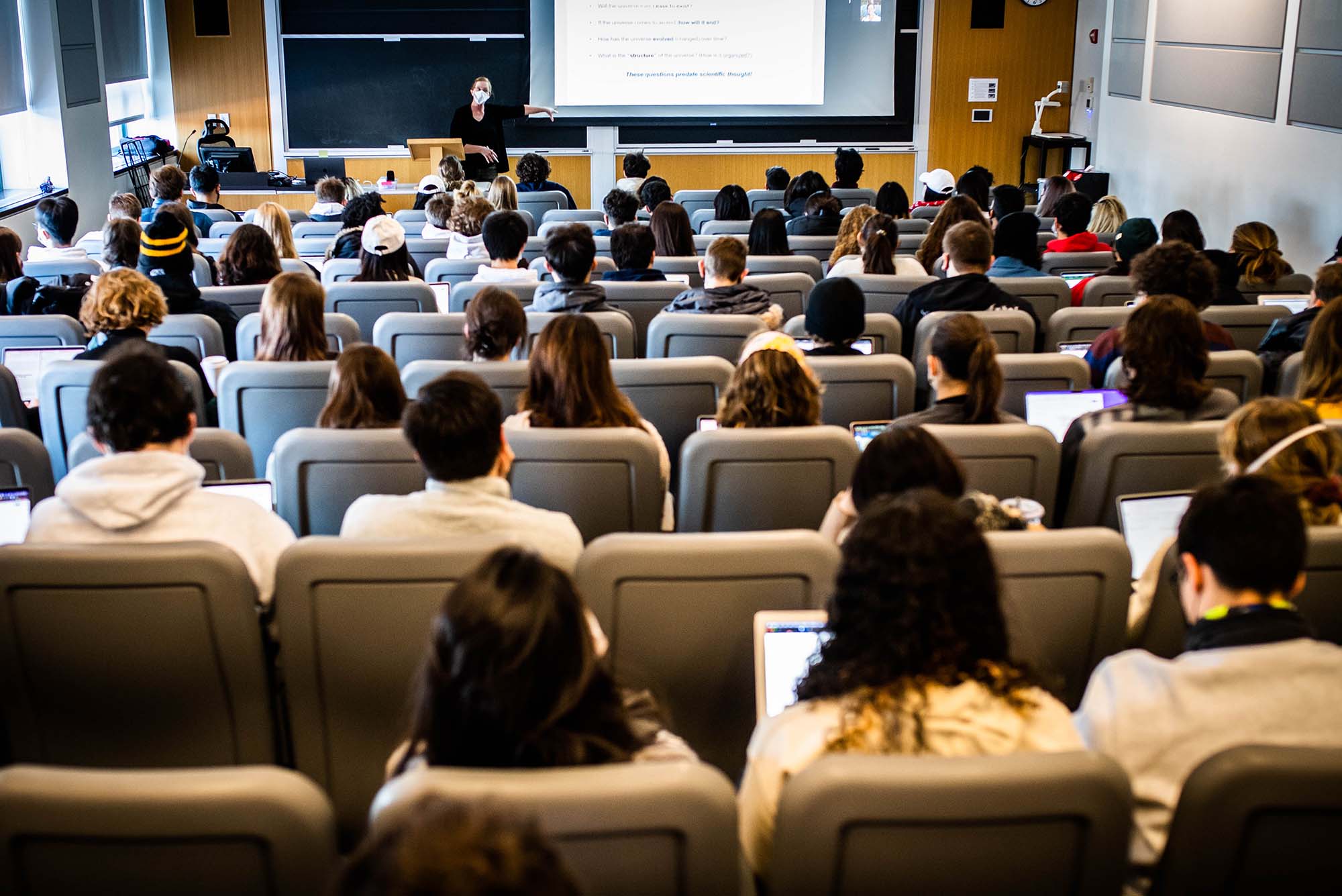 Photo of Astronomy professor, Tereasa Brainerd, at the front of the lecture hall to the left of a projector screen. Students fill the rows of her cosmology class on January 21, 2022.