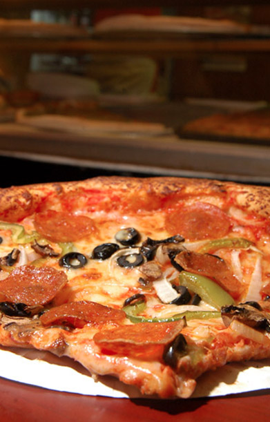 Closeup photo of a supreme pizza pie with olive, peppers, and pepperoni. A blurry pizza oven can be seen in the background.