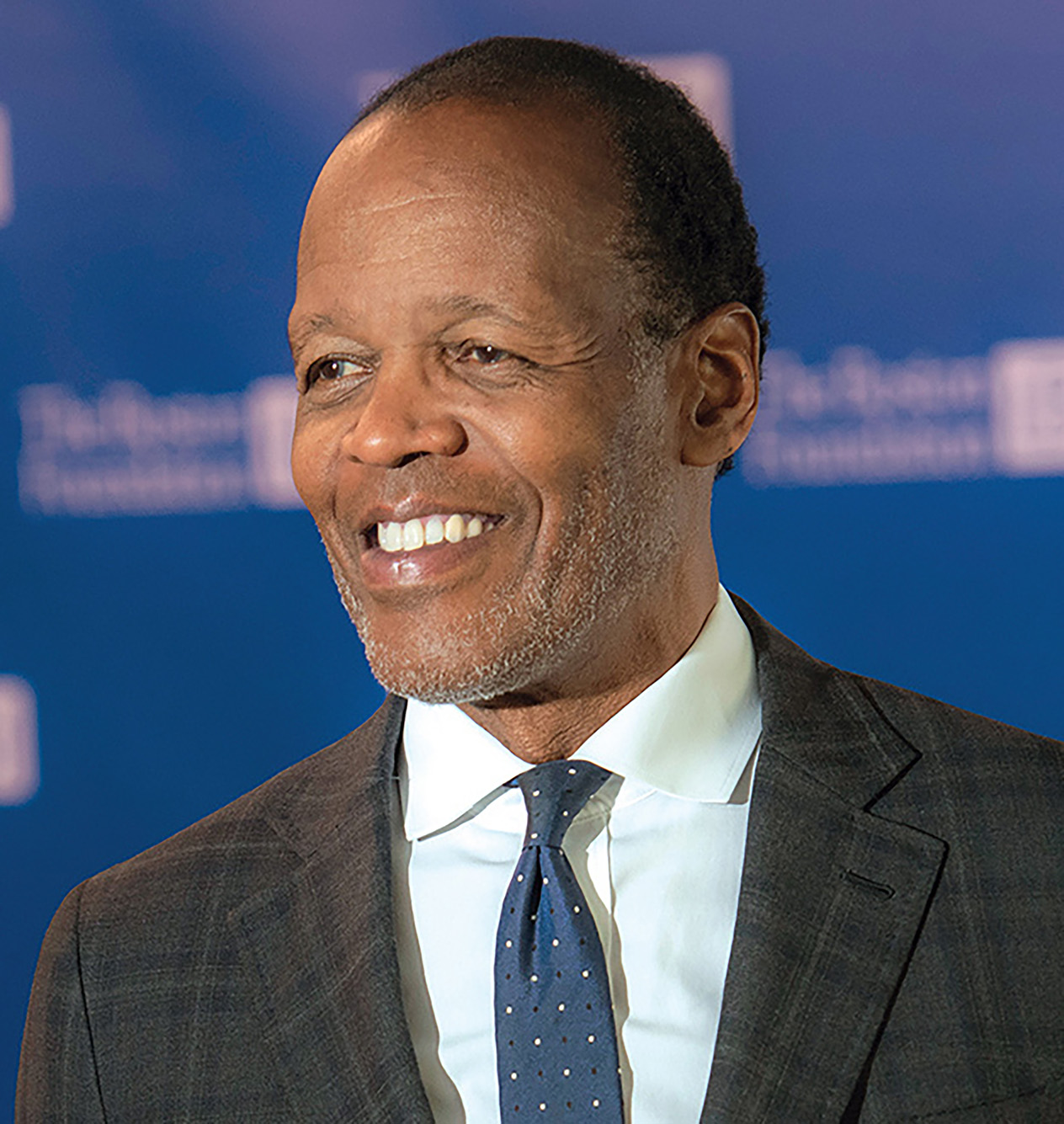 photo of Lee Pelton, a black man with gray stubble wearing a black suit, white shirt, and tie. He looks to the left in front of a darer blue background.