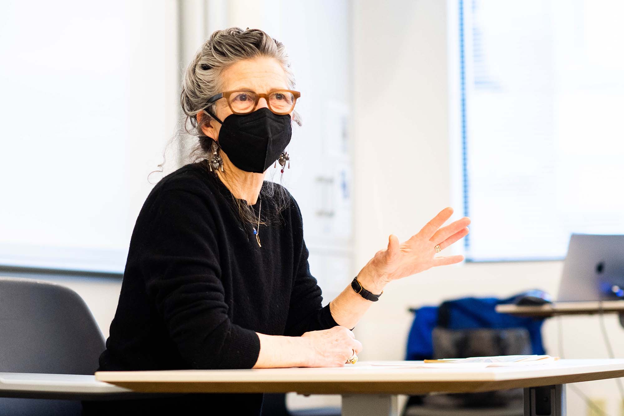 Photo of Jody Adams, wearing a black face mask, black shirt, and brownish glasses, as she sits at the front of a classroom. She moves her hand as she speaks. Her salt-and-pepper hair is pulled back.