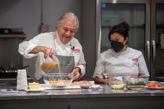 Photo of Jacques Pépin (Hon.’11), assisted by Lisa Falso-Doherty, as he pours stock into the bread and spinach stuffing for chicken ballottine in the newly renovated Groce-Pépin Culinary Innovation Lab kitchen. Pépin wears a gray Pépin foundation apron and chef jacket with BU logo, and Falso-Doherty also wears a chef jacket with the BU logo and wears a black mask. Bowls of ingredients rest on the counter before them.