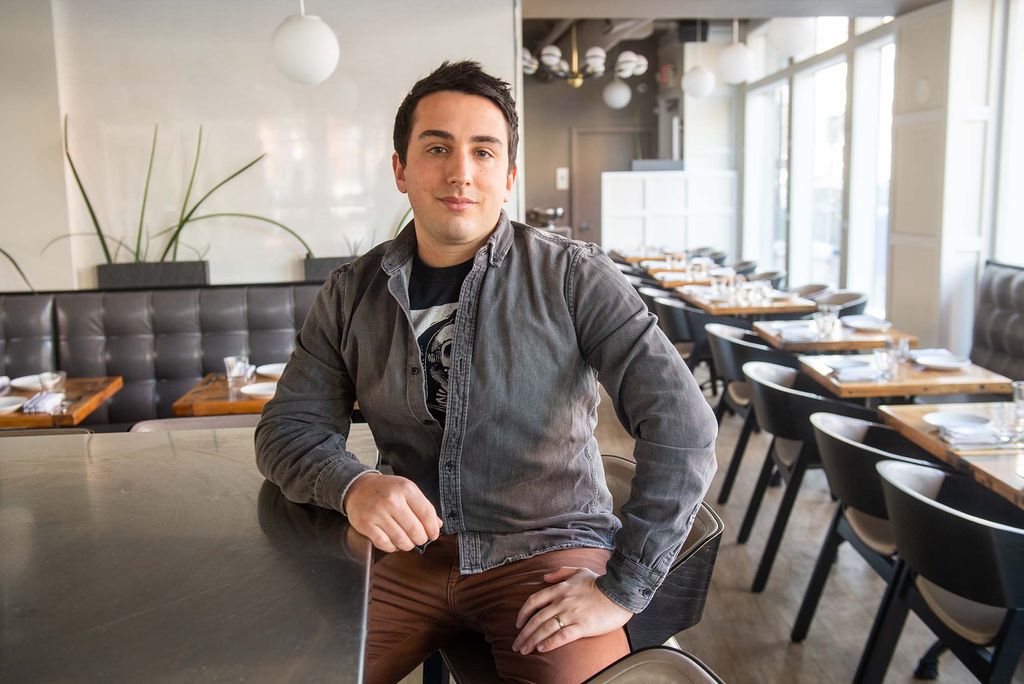 Photo of Seth Gerber, a young light-skinned man with short brown hair sitting at the bar of his South Boston restaurant Mida, wearing a washed gray jacket and brown pants. The wooden restaurant tables are set and have gray chairs or gray booth seats.