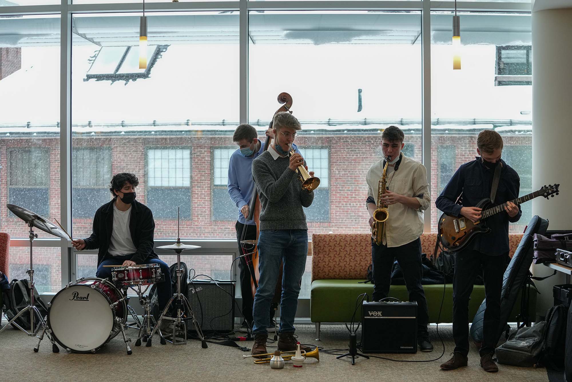 Photo of BU's Jazz Band performing on Feb 13 at Campus Center and Student Residence building on Fenway Campus. Five young men play, some with masks on. There is a drummer, bassists, trumpeter, saxophone and jazz guitar.