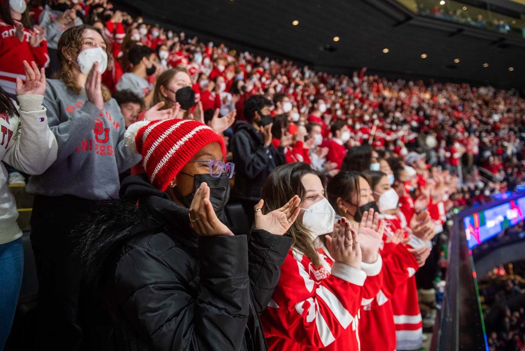 Photo of a young student of color clapping her hands during the beanpot game. She wears a black puffy, glasses, face mask, and a BU hat. Other fans cheer around her.