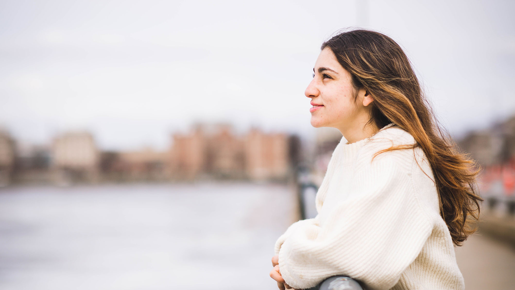 Photo of Sara Tavakoli (SDM’25). She is seen in profile, and leans on a railing along the Charles River looking out towards the water. She is light-skinned, has brown hair with highlights and wears a white sweater.