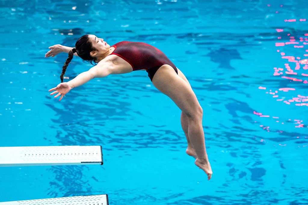Photo of Emily Balicanta (Sargent’22) as she impressively springs off of a white diving board in the pool below. She is a young woman, who wears a red swim suit and has her dark hair braided in a pony tail.