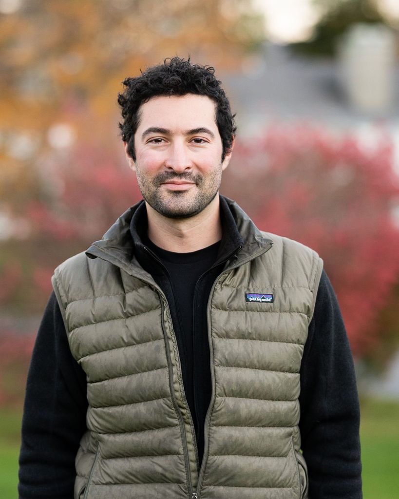 Photo of Elias Weiss Friedman, a white man with dark black hair and short beard. He wears a black long sleeve with a green patagonia vest.