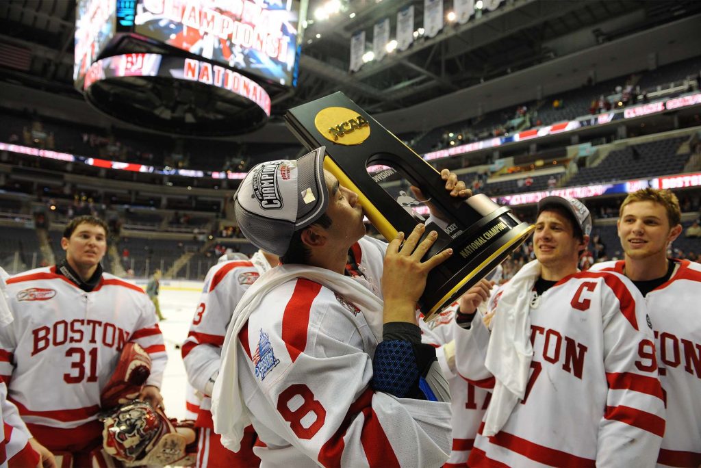 Photo of Brandon Yip (CAS’09), a member of the 2008-09 NCAA championship-winning Terrier squad, kissing the NCAA trophy and wearing a champions hat. He stands center rink in a large stadium and his smiling team mates are seen around him.