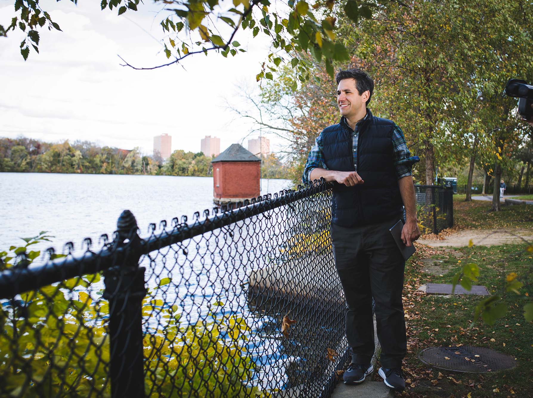 photo of Andrew Robichaud, a white man wearing a blue plaid shirt and a navy puffer vest and dark blue jeans, stands next to a fence and looks out over a river