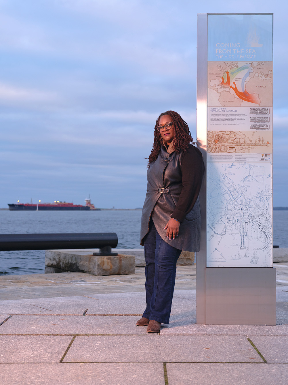 Photo of Deborah Douglas, a dark-skinned Black woman with long dreads wearing stylish glasses and a vegan leather vest, long-sleeved black shirt, and jeans leans back against a a tourist map pillar. A beautiful purple sunset amidst a cloudy sky and large body of water is seen behind her.