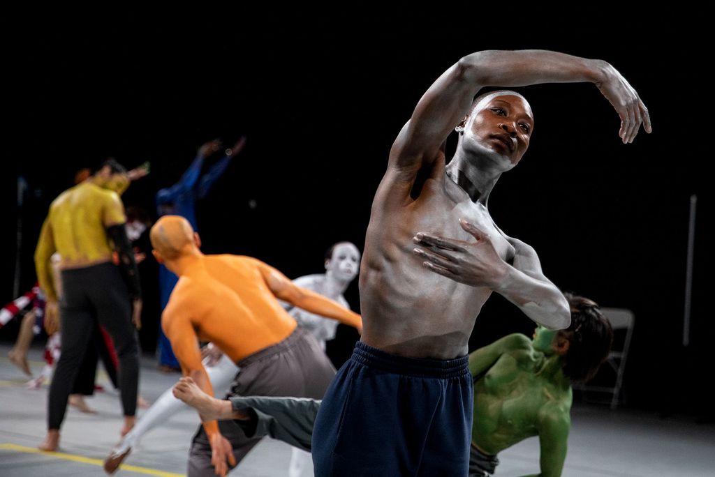 Photo of Dancers in Jones’ Curriculum. In the foreground at right, a young Black man with white paint on his chest curves his right arm over his head gracefully. In the background, other dancers, painted orange, green and white, dance on stage.