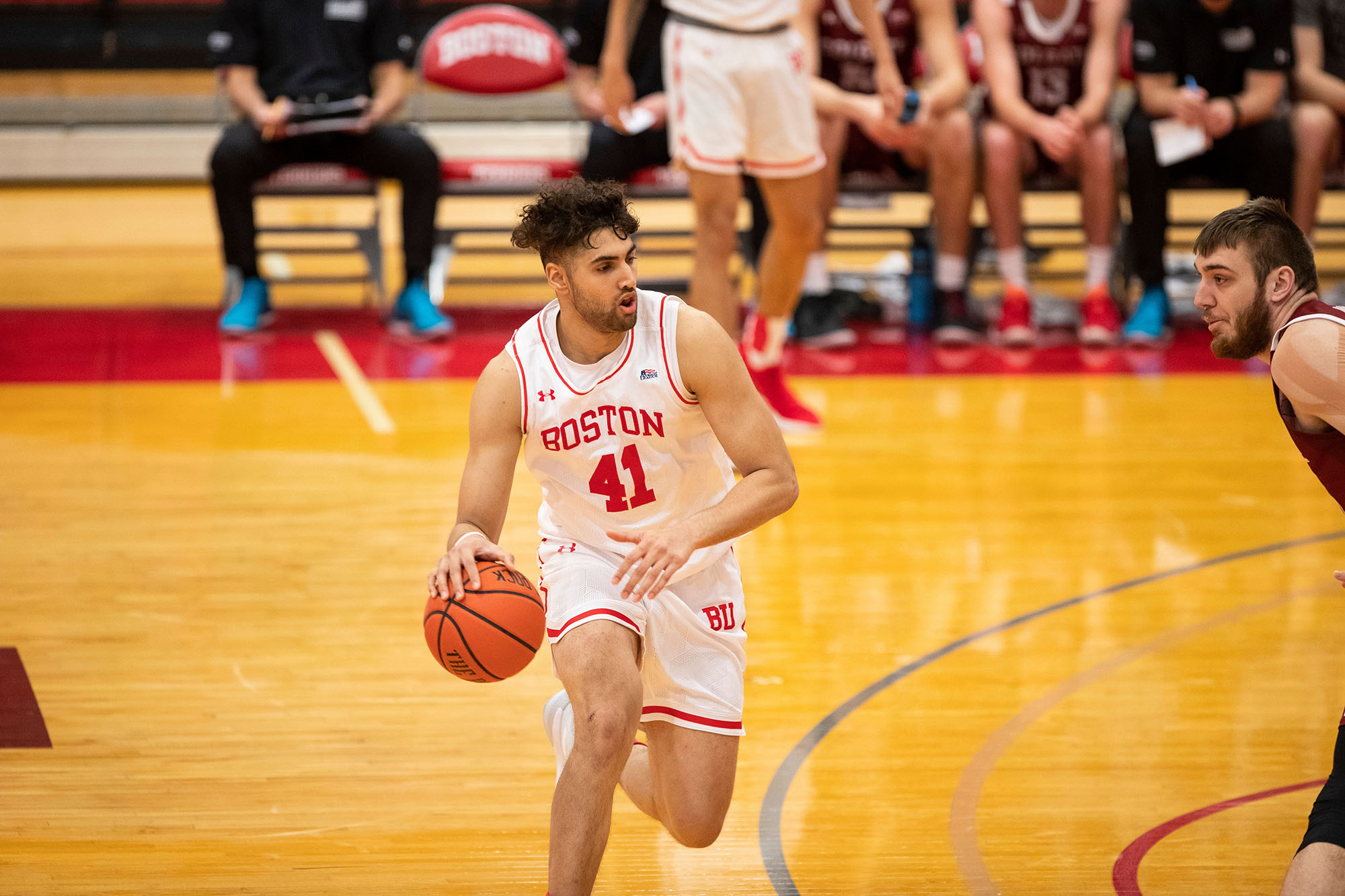 Photo of Mathon mid-game as he dribbles down a basketball court. A tan young adult with brown curly hair wears a white and red jersey as he makes his way down court. Blurry background shows other members of BU men's basketball team sitting on the bench.