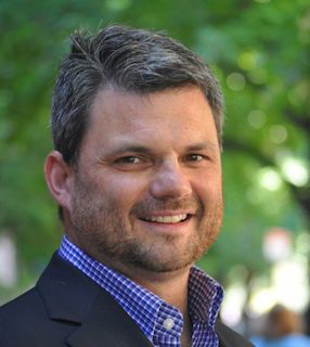 Headshot photo of Kevin Gallagher. A white man with graying hair looks and smiles at the camera. A blurry, green tree is seen in the background.