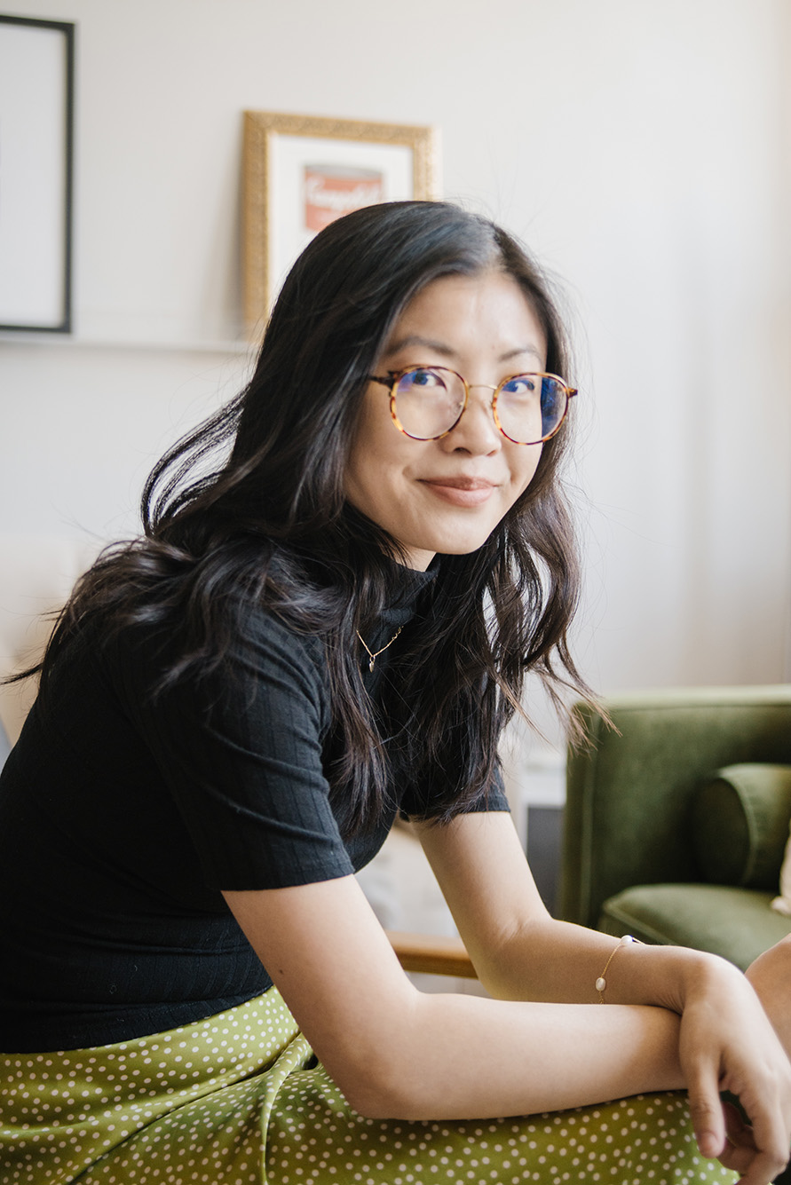 Photo of Weike Wang (GRS’15), a young Asian American woman with shoulder length brown hair and turtle glass. She wears a black blouse and spotted green and yellow skirt and sits with her hands folded as she slightly smiles.