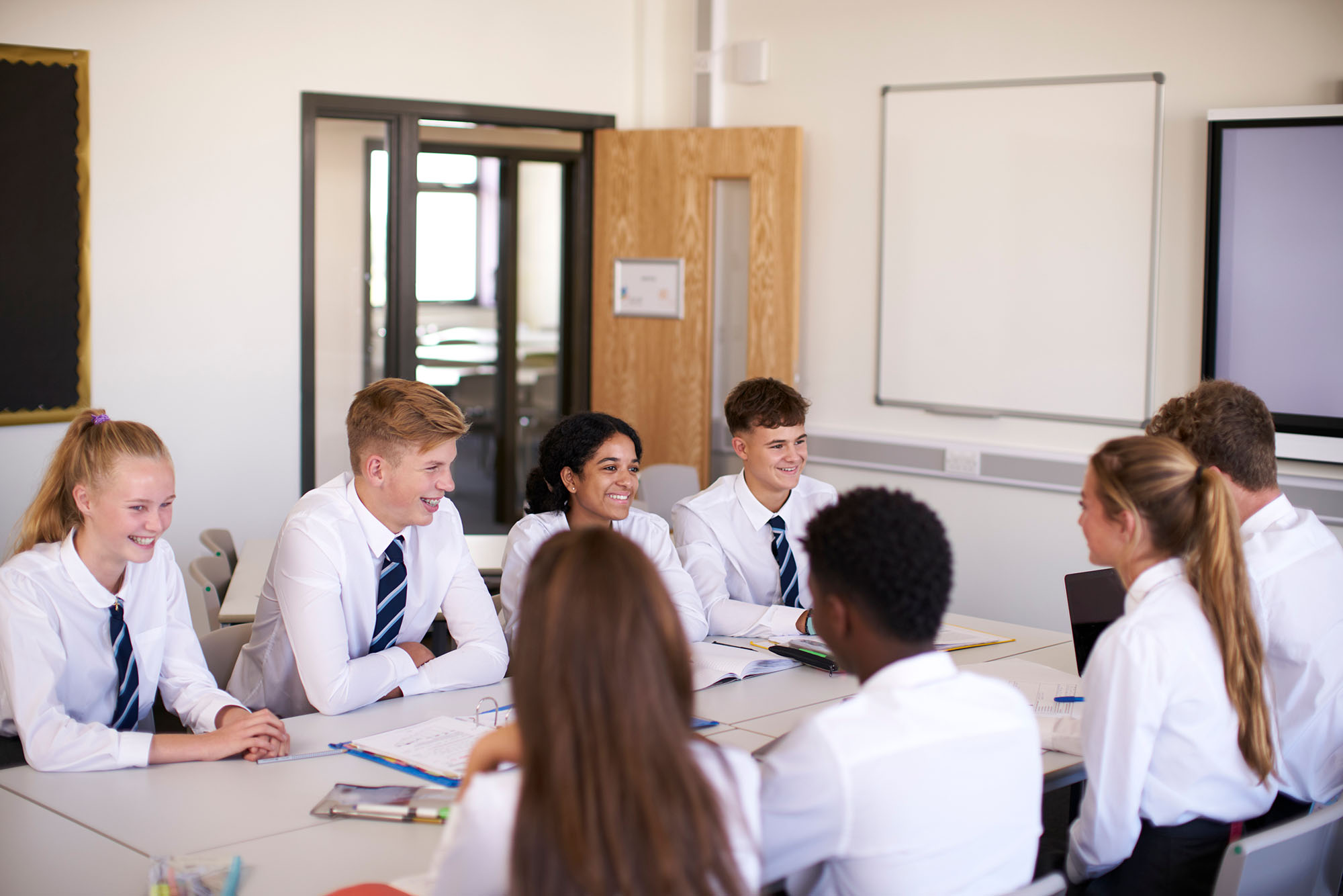 Photo of students wearing a private school uniform of white collared shirts, blues ties, and blue skirts and pants. They all sit facing each other in a bright classroom.