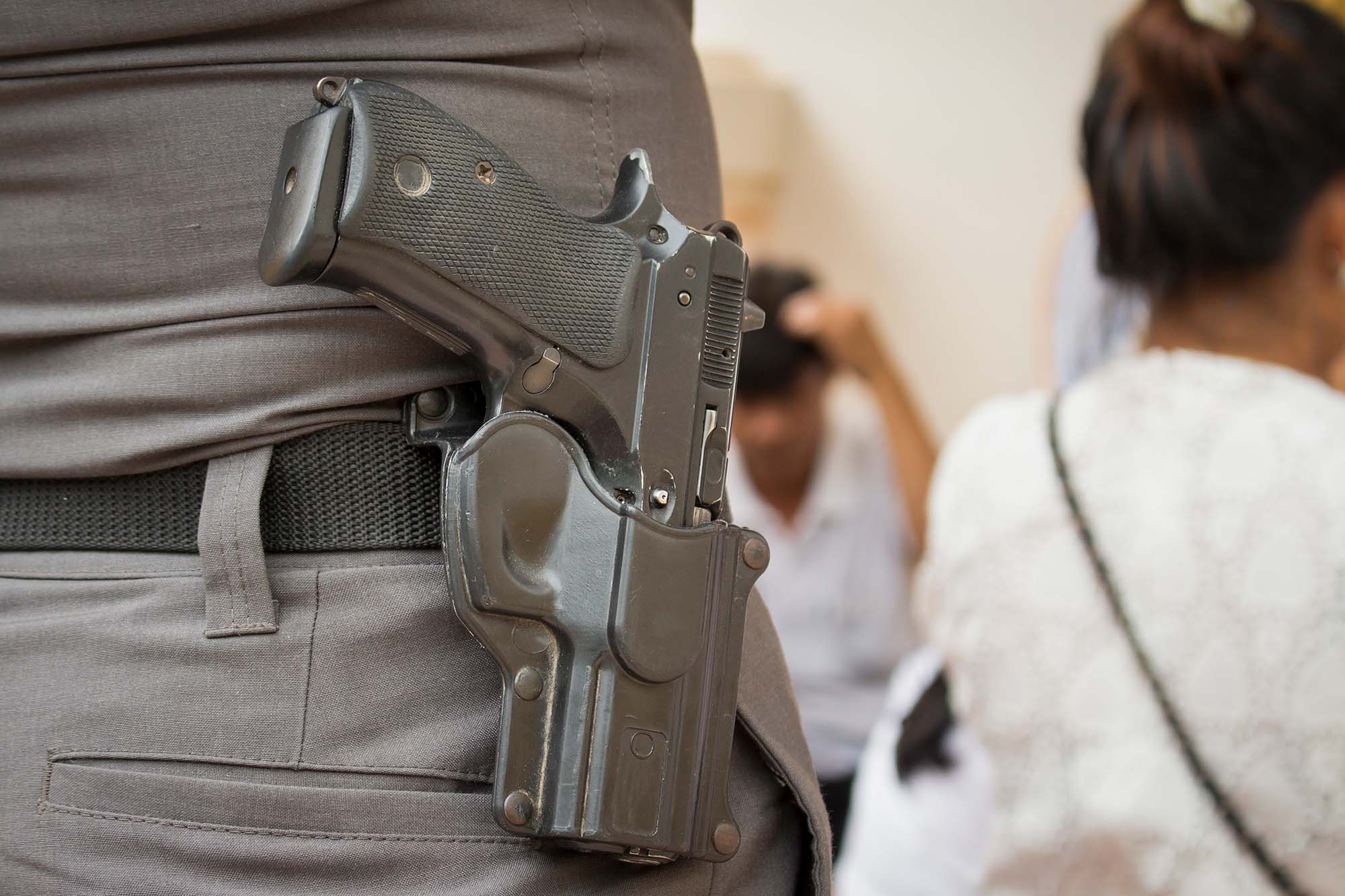 Closeup photo of someone with a gun in their holster. The holster is attached to their pants. Two blurry people can be seen in the background at what looks to be a cafe.