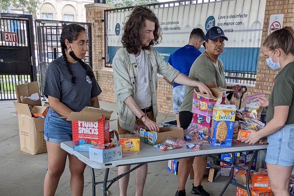 Photo: A few volunteers packing care packages with the nonprofit Team Brownsville at the Brownsville bus station. A plastic table is setup with various packages of non-perishables items are on and around the table.