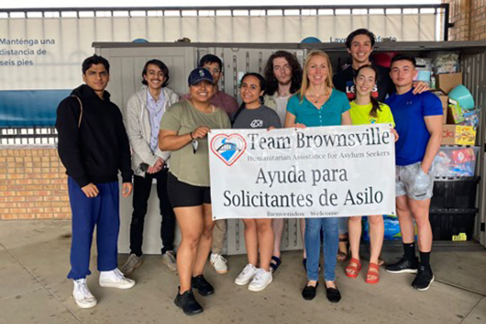 Photo: The border studies group stands in courtyard and poses as they hold up a sign that reads "Team Brownsville. Ayuda para Solicitantes de Asilo"