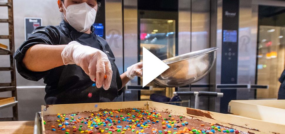 Carmen Quijada adds sprinkles to a batch of brownies. There is a white play button overlayed on top of the image