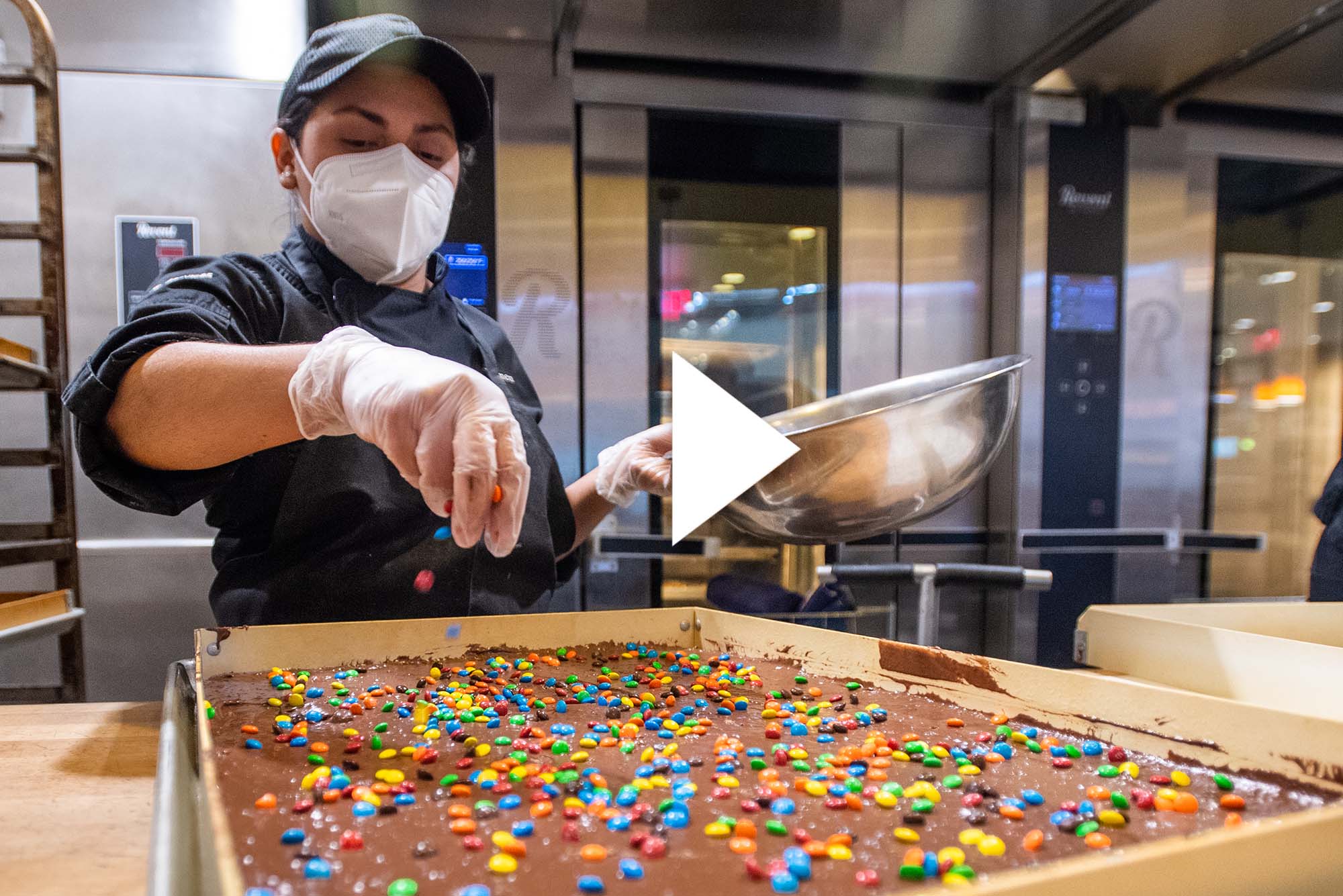 Carmen Quijada adds sprinkles to a batch of brownies. There is a white play button overlayed on top of the image