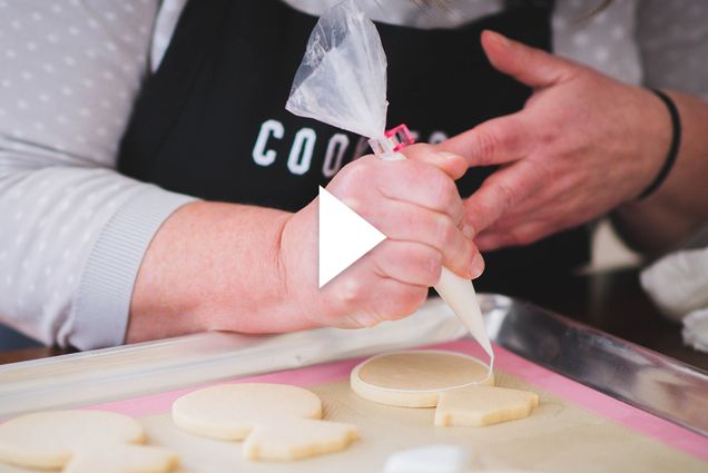 someone pipes frosting out of a bag onto white cookies on a baking sheet. There is a white play button overlayed in the center of the image