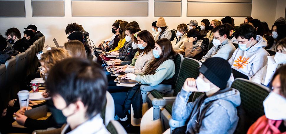 Photo of a class of masked BU students sitting in a lecture hall. They wear a variety of winter coats and outfits as the look to the left towards the front of the room.