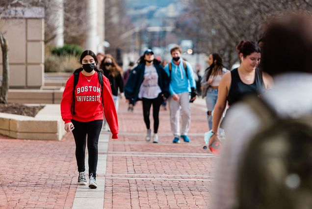 Stock photo of BU West campus. photo is focused on a masked young woman in a bright red hoodie that reads "Boston University" as she walks down Comm Ave. Various masked and unmasked people walk up and down the cobbled sidewalk around her.