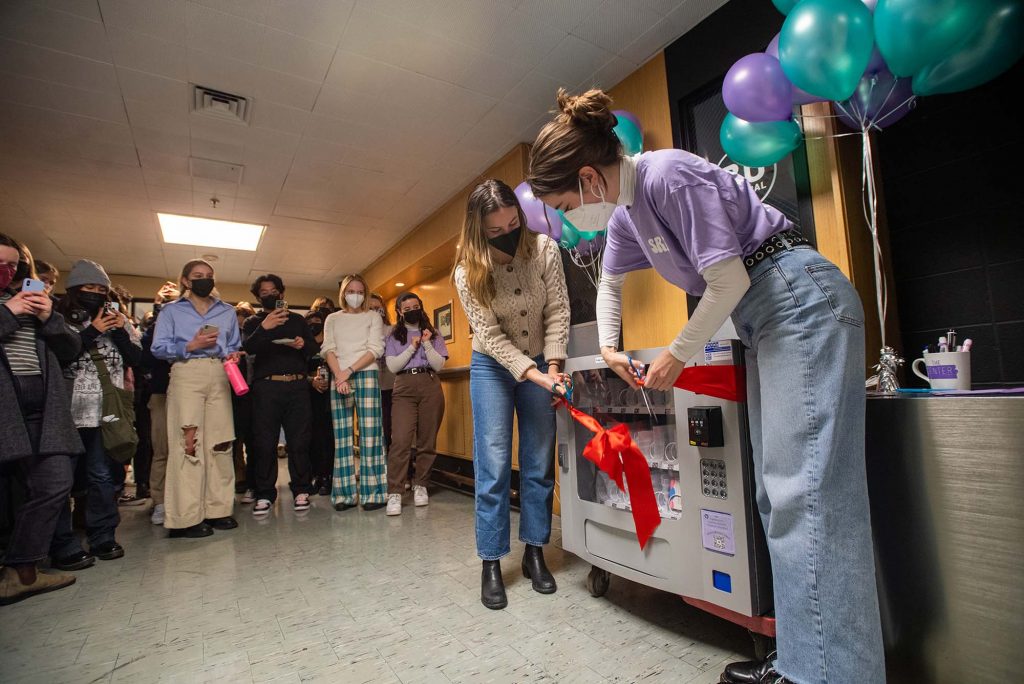 Photo of Students for Reproductive Freedom co-presidents Molly Baker (CAS’22), left, and Charlotte Beatty (CAS’22) cutting the ribbon on the new vending machine during a launch party in the GSU. Two masked students bend and cut a large red ribbon tied around the machine as masked onlookers circle around and clap.