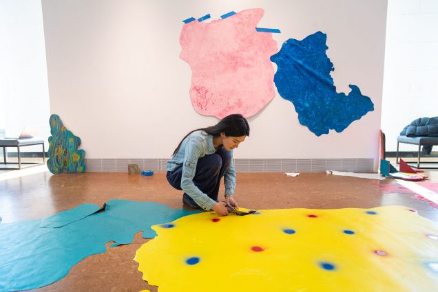 Photo of Moxin “Sunny” Chen (CFA’22) installing her vinyl-and-paint collage piece, “Yesterday Is Passed, Tomorrow Has Yet to Come". A young, Asian woman bends down and adjusts a large yellow blob that features near many other various colored blobs in the exhibit.