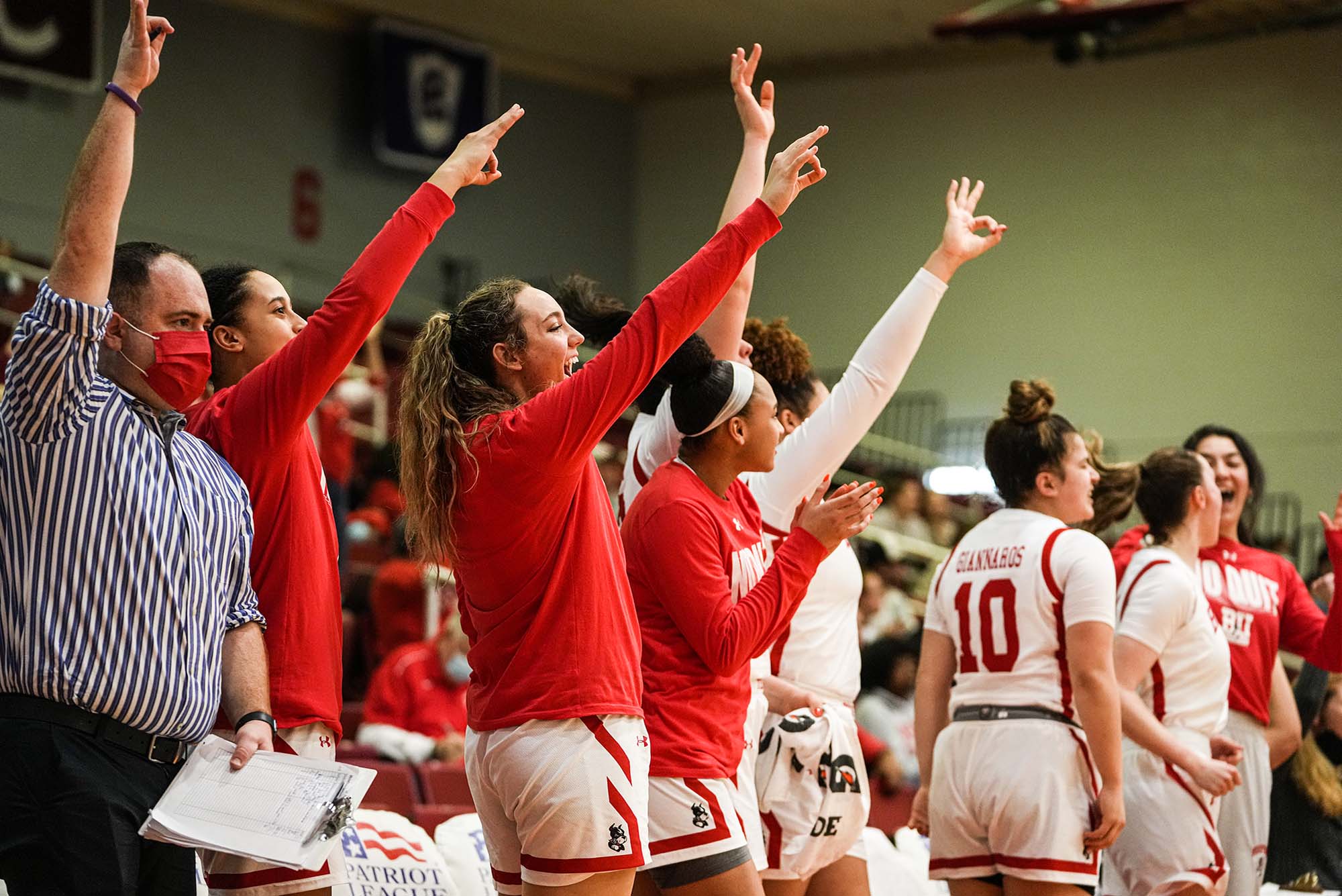 Photo of the members of BU Women's Basketball team from the bench cheering. 4 women wearing red long-sleeved shirts hold their arms up and hold their fingers in a dog formation to cheer. A masked ref is seen on the left raising his arm.