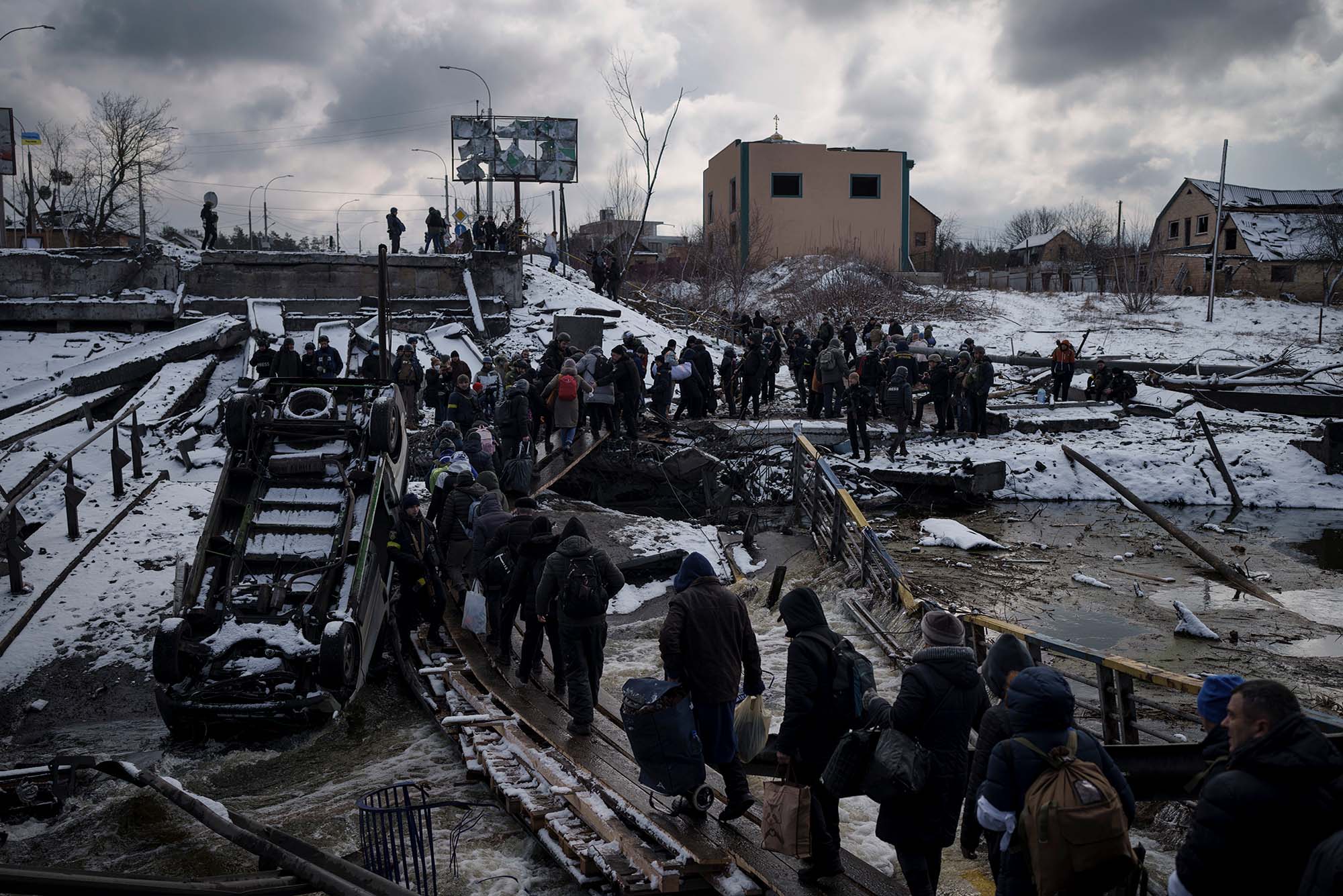 Photo of Ukrainian citizens crossing an improvised path under a destroyed bridge while fleeing Irpin on March 8. Photo shows a line of people wearing dark clothes on a dark snowy day as they follow a path near a destroyed bridge.