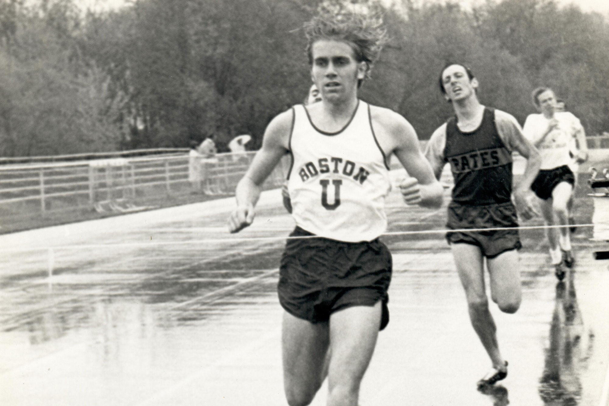 Black and white photo of Tom Beatty (CAS’72) during a historic track meet. He wears a BU jersey and his hair bounces as he runs ahead of two competitors on a track.