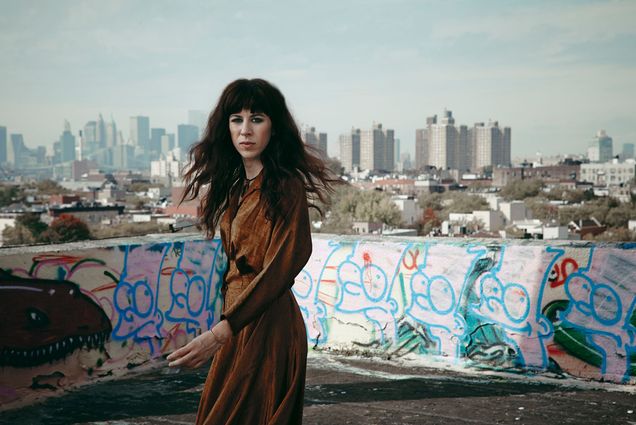 Photo of Missy Mazzoli, a white women with long brown hair. She wears a brown dress and is caught mid-turn on a roof with artistic graffiti covering it's walls. A city scene can be seen in the distance.